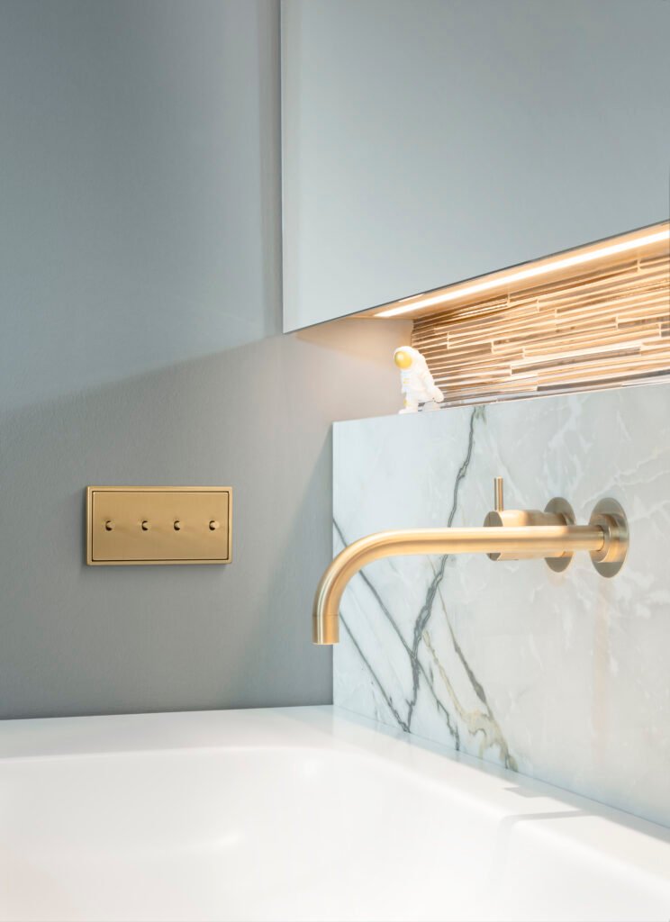 Išskirtiniai JUNG jungikliai – pirmas pasirinkimas prabangaus būsto interjerui 3 Minimalaus stiliaus interjero dizainas. Vonios prastuvė su auksinės spalvos akcentais ir jungikliu melsvame fone.